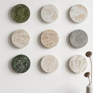 Op de afbeelding: Negen cirkelvormige stenen wandplaten gerangschikt in een raster, met diverse natuurlijke texturen en kleuren, waaronder bosgroene, beige, crèmekleurige en grijze marmerpatronen. Een witte vaas met gedroogde lotuspeulen staat op een donkerhouten oppervlak.