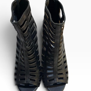 May include: A pair of black ankle boots featuring a cage-style design with multiple horizontal cutouts across the upper. These open-toe boots have a high-cut silhouette and include a vertical zipper closure at the back of each heel for secure fastening.