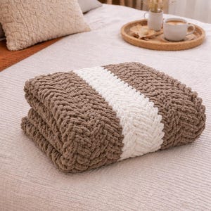 Könnte beinhalten: Eine gefaltete, grob gestrickte Wohndecke mit einem breiten, weißen Mittelstreifen, der von zwei breiteren braunen Abschnitten flankiert wird. Die Decke besteht aus weichem, strukturiertem Garn mit einem sichtbaren Fischgrätenmuster.