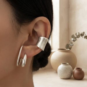 May include: A silver ear cuff with a wide, smooth band sits on the upper cartilage, while two polished silver hoop earrings of varying sizes are worn on the earlobe, creating a layered metallic look on the ear.