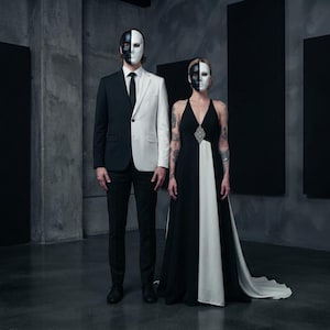 May include: A two-tone suit jacket featuring a black left side and white right side, paired with black trousers and a black tie. Beside it, a floor-length gown with a black bodice, white skirt panel, and a central silver crystal embellishment.