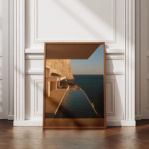 May include: A rectangular framed wall art print featuring a photograph of a modern architectural structure with a terrace and an infinity pool overlooking a deep blue sea and a large rocky cliff face under a clear sky during the golden hour.