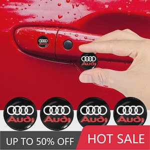 May include: A set of circular black adhesive emblems featuring the white Audi logo and red text. One emblem is being applied to a red car door handle, while four identical emblems are displayed in a row below against a white background.