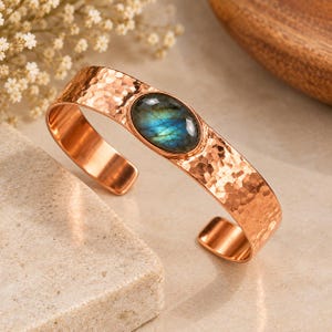 May include: A copper cuff bracelet featuring a hammered, textured surface and a central oval-shaped labradorite gemstone. The stone displays iridescent blue and green flashes set within a smooth, polished copper bezel frame.