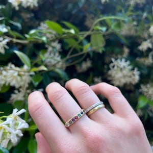 May include: A gold ring featuring a row of five small, round, multi-colored gemstones including green, red, and blue stones, paired with two additional thin gold bands, one of which is set with tiny clear stones and the other is a plain gold band.