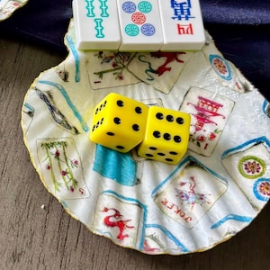 May include: A decorative scallop shell dish featuring a decoupage pattern of mahjong tiles, holding three white mahjong tiles with green, red, and blue symbols and two bright yellow six-sided dice with black pips resting in the center.