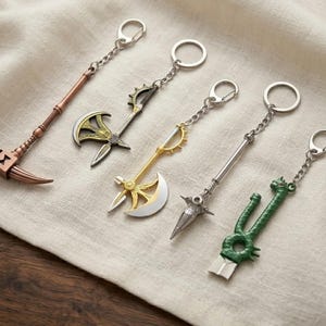 May include: Five metal keychains shaped like fantasy weapons, including a copper-coloured scythe, two axes with gold and silver details, a silver spear-like weapon, and a green dragon-themed blade, all attached to silver rings.