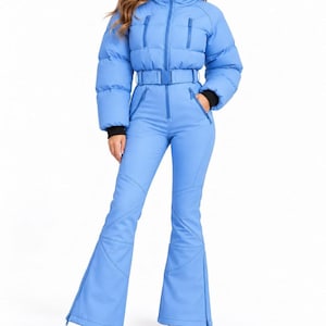 Può includere: Una tuta da sci intera color azzurro con corpetto imbottito, collo alto, chiusura frontale con zip e due tasche pettorali con cerniera. La vita è dotata di una cintura abbinata e i pantaloni presentano un taglio a zampa con polsini neri.