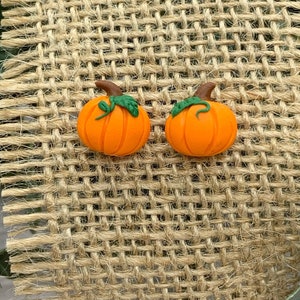 May include: A pair of orange pumpkin earrings with green stems and leaves. The earrings are displayed on a brown burlap background.