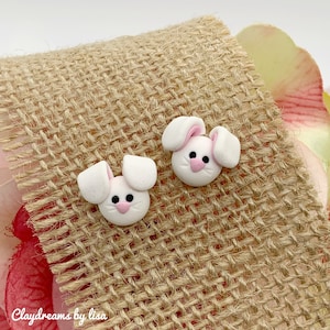 May include: A pair of white bunny rabbit stud earrings with pink ears and noses. The earrings are on a burlap background.