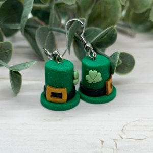 May include: A pair of green leprechaun hat earrings with a gold buckle and a shamrock on each hat. The earrings are made of polymer clay and have silver hooks.