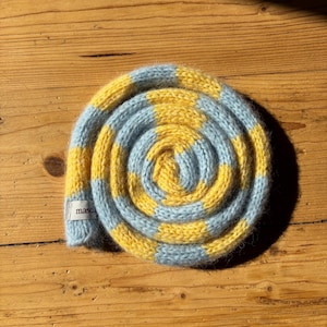 Könnte beinhalten: Ein aufgerollter Strickschal mit abwechselnden horizontalen Streifen in Hellblau und Gelb. Die weiche, flauschige Textur des Garns ist in der gesamten Spirale sichtbar. Ein kleines weißes Stoffetikett mit dem schwarzen Text masci ist am Ende befestigt.