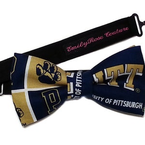 May include: A navy blue and gold bow tie with the University of Pittsburgh logo and a paw print. The bow tie is attached to a black adjustable strap with the text "Emily Rose Couture" in pink.