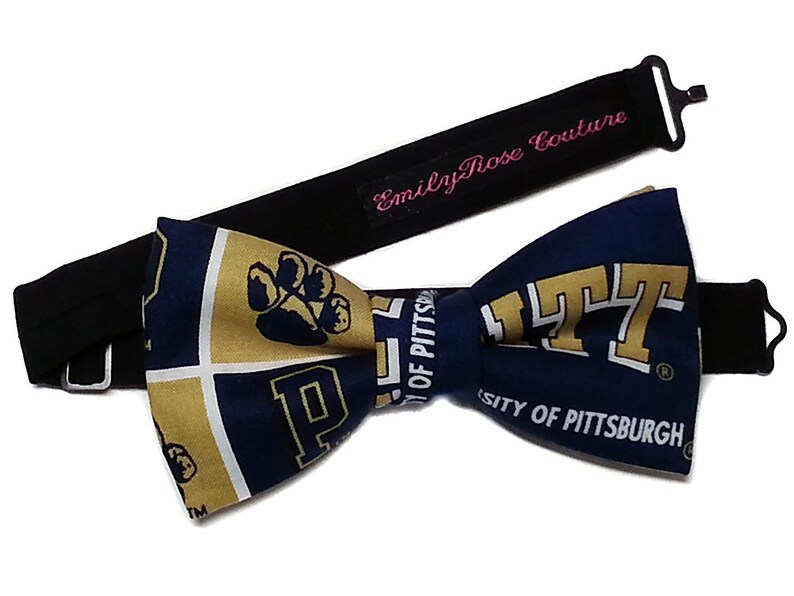 May include: A navy blue and gold bow tie with the University of Pittsburgh logo and a paw print. The bow tie is attached to a black adjustable strap with the text "Emily Rose Couture" in pink.