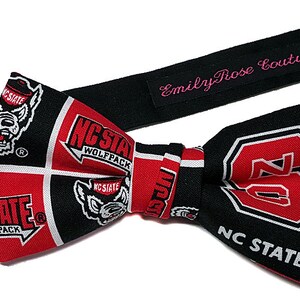 May include: A black bow tie with a red and white pattern featuring the NC State Wolfpack logo. The bow tie is attached to a black adjustable strap with the text "Emily Rose Couture" in pink.
