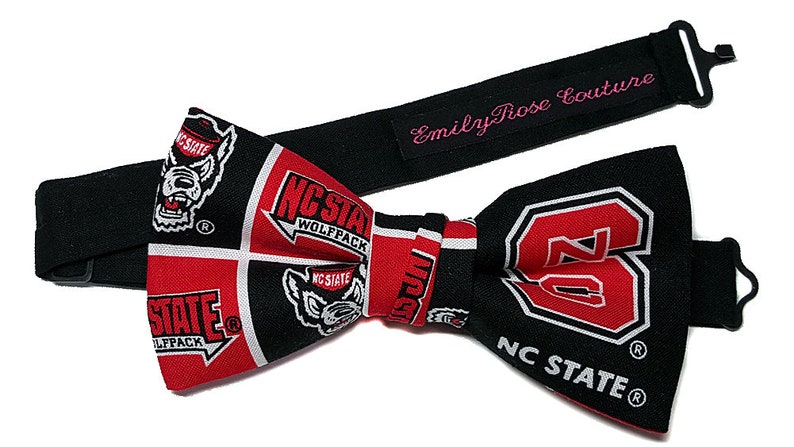 May include: A black bow tie with a red and white pattern featuring the NC State Wolfpack logo. The bow tie is attached to a black adjustable strap with the text "Emily Rose Couture" in pink.