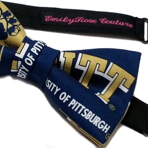 May include: A blue and gold bow tie with the University of Pittsburgh logo. The bow tie is made of fabric and has a black adjustable strap with a metal clasp.