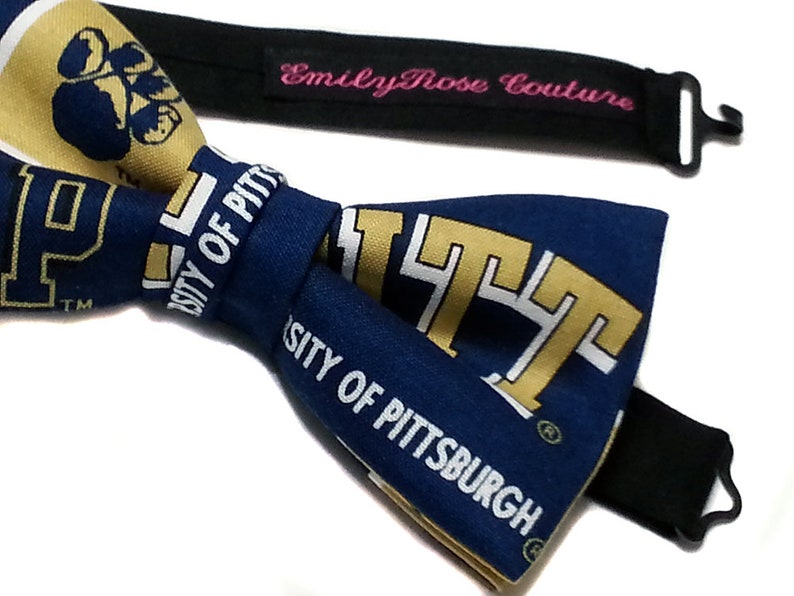May include: A blue and gold bow tie with the University of Pittsburgh logo. The bow tie is made of fabric and has a black adjustable strap with a metal clasp.