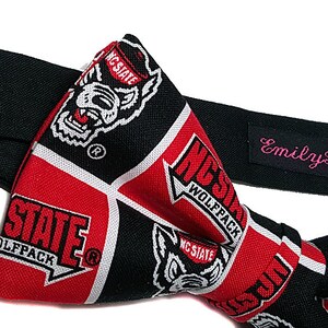 May include: A black bow tie with a red and white pattern featuring the NC State Wolfpack logo. The bow tie is made by Emily Rose Couture.