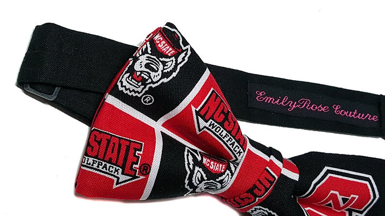 May include: A black bow tie with a red and white pattern featuring the NC State Wolfpack logo. The bow tie is made by Emily Rose Couture.