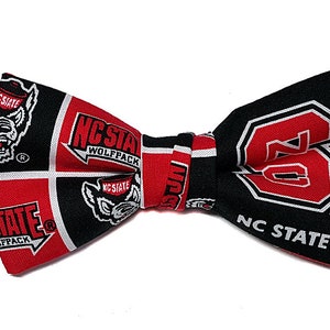 May include: A black bow tie with an adjustable strap. The bow tie is made of fabric with a red and black pattern featuring the NC State Wolfpack logo and the text "NC State Wolfpack".
