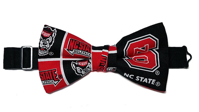 May include: A black bow tie with an adjustable strap. The bow tie is made of fabric with a red and black pattern featuring the NC State Wolfpack logo and the text "NC State Wolfpack".