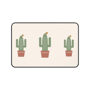 Puede incluir: Una alfombra rectangular con fondo color crema y borde negro. El diseño presenta tres cactus verdes en macetas de color terracota, cada uno coronado con un pequeño círculo amarillo, dispuestos horizontalmente en el centro de la alfombra.