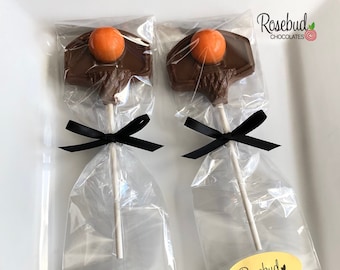 以下が含まれることがあります： オレンジ色のバスケットボールが乗ったチョコレートのバスケットボールロリポップが2本、透明なセロファン袋に黒のリボンで結ばれ、ゴールドのラベルには「Rosebud Chocolates」と書いてあります。