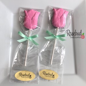 May include: Two pink chocolate rosebud lollipops wrapped in clear cellophane bags with green ribbon bows. Each lollipop has a white stick and a yellow oval sticker with the text "Rosebud Chocolates".