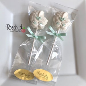 May include: Two white chocolate baby carriage lollipops with green accents, individually wrapped in clear cellophane bags with a green ribbon and a gold sticker that says "Rosebud Chocolates".