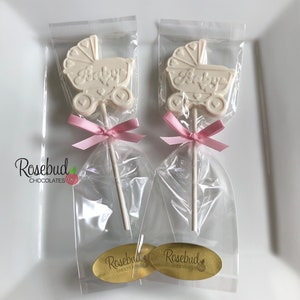 May include: Two white chocolate lollipops shaped like baby strollers with the word "Baby" on them. The lollipops are wrapped in clear cellophane bags with pink ribbons. The bags have gold stickers with the text "Rosebud Chocolates" on them.