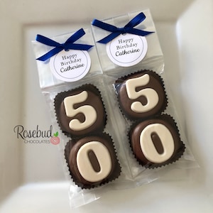 May include: Two clear cellophane bags with blue ribbon bows, each containing two chocolate-covered cookies. The cookies are decorated with white icing and the numbers 5 and 0. The bags have a white tag with the text "Happy Birthday Catherine".