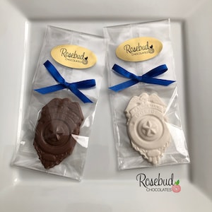 May include: Two chocolate police badges in clear cellophane bags tied with blue ribbon. The badges are in the shape of a shield with a star in the center. One badge is brown and the other is white. The bags have a gold label that says "Rosebud Chocolates".