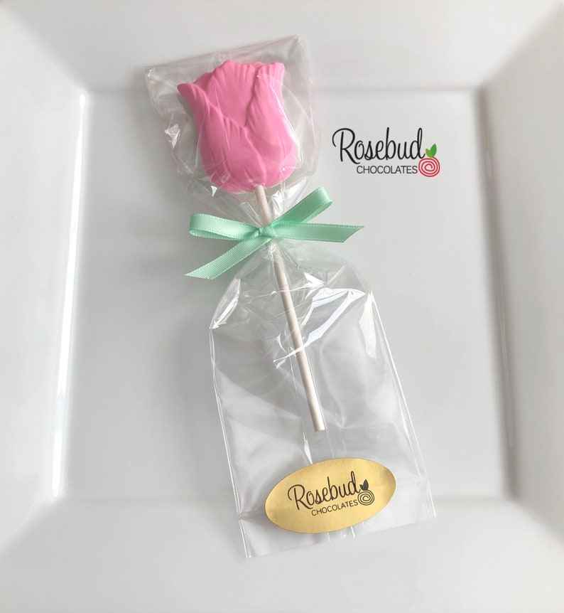 May include: A pink chocolate rose lollipop wrapped in clear cellophane with a green ribbon and a gold label that says "Rosebud Chocolates".