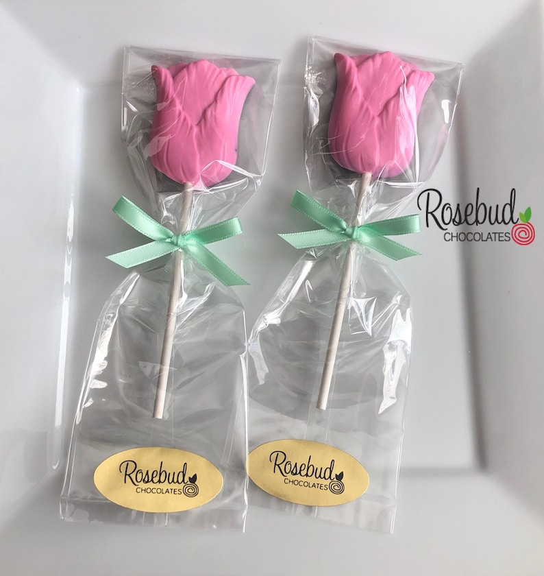 May include: Two pink chocolate rosebud lollipops wrapped in clear cellophane bags with green ribbon bows. Each lollipop has a white stick and a yellow oval sticker with the text "Rosebud Chocolates".