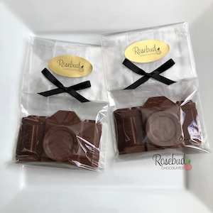 May include: Two chocolate camera favors in clear cellophane bags tied with black ribbon. Each bag has a gold oval sticker with the text "Rosebud Chocolates".