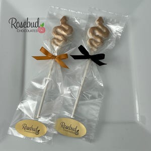 May include: Two gold chocolate lollipops shaped like a snake, wrapped in clear cellophane bags with a gold and black ribbon. The bags have a label that says "Rosebud Chocolates".