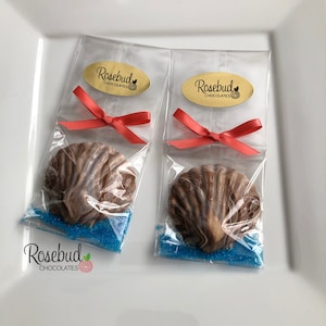 May include: Two clear cellophane bags with red ribbon bows, each containing a chocolate seashell and blue sprinkles. The bags have gold oval stickers with the text "Rosebud Chocolates" and a small rose graphic.
