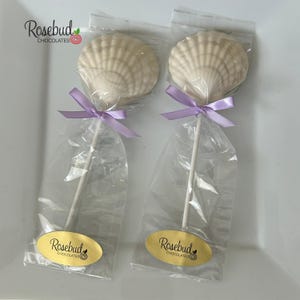May include: Two white chocolate seashell lollipops individually wrapped in clear plastic bags. Each lollipop features a light purple ribbon tied around the white stick and an oval gold sticker with the text Rosebud Chocolates at the base.