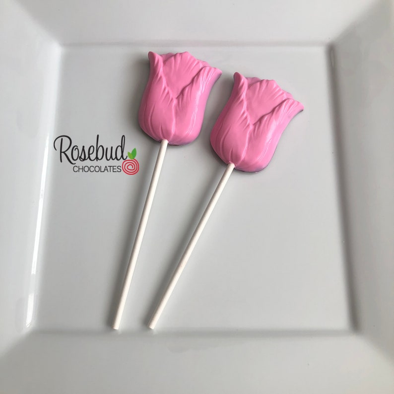 May include: Two pink chocolate tulip lollipops on a white plate. The lollipops are on white sticks.