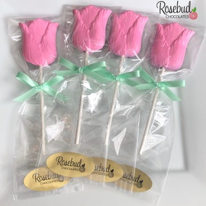 May include: Four pink chocolate tulip lollipops wrapped in clear cellophane bags with green ribbon bows. Each lollipop has a white stick and a gold sticker with the text "Rosebud Chocolates" and a small circle with a rosebud design.