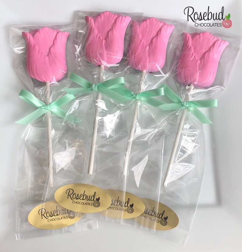May include: Four pink chocolate tulip lollipops wrapped in clear cellophane bags with green ribbon bows. Each lollipop has a white stick and a gold sticker with the text "Rosebud Chocolates" and a small circle with a rosebud design.
