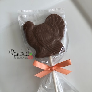 May include: A chocolate turkey lollipop wrapped in clear cellophane with an orange ribbon. The lollipop is on a white stick and has the text "Rosebud Chocolates" on the packaging.
