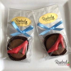 May include: Two clear cellophane bags tied with blue ribbon, each containing a chocolate favor with a red high heel shoe design. The bags have a gold oval sticker with the text "Rosebud Chocolates".