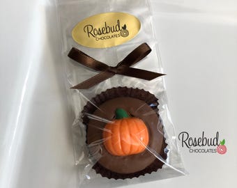 May include: A round chocolate treat topped with an orange pumpkin decoration, sitting in a dark brown paper liner inside a clear plastic bag. The bag is tied with a brown ribbon and features a gold oval label reading Rosebud Chocolates.