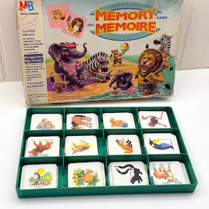 Puede incluir: Una caja de juego de memoria Milton Bradley con ilustraciones de animales y el texto Memory Jeu Mémoire, acompañada de una bandeja de plástico verde que contiene doce pilas de tarjetas blancas cuadradas con dibujos de animales coloridos.