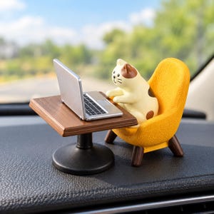 May include: A miniature figurine of a cream and brown cat sitting in a textured yellow armchair at a small brown table with a silver laptop. The cat has brown patches on its head and back, and the table features a dark brown circular base.