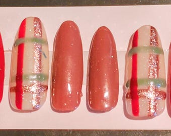 May include: A set of ten press-on nails featuring a variety of designs including solid sage green, solid bright red, solid muted rose, and a cream base with a red and green plaid pattern accented by thin lines of shimmering copper glitter.