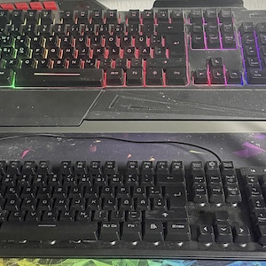 Könnte beinhalten: Ein schwarzer Computer-Tastaturständer mit einer oberen Tastatur mit mehrfarbiger RGB-Hintergrundbeleuchtung und einer unteren Tastatur mit schwarzen Tasten. Beide Tastaturen verfügen über ein QWERTZ-Layout mit einem Ziffernblock.