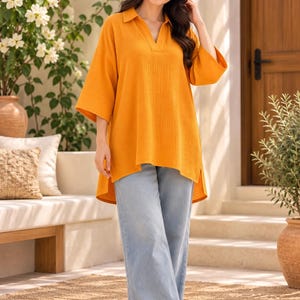 May include: A mustard yellow tunic top with a collared V-neckline and three-quarter length sleeves, paired with light blue wide-leg denim jeans and white slide sandals. The tunic features a textured fabric and a high-low hemline.